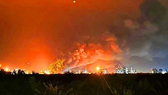 Nighttime scene of multiple industrial refinery facilities engulfed in large orange flames and thick black smoke billowing upward with scattered bright lights from surrounding structures and towers visible against a dark sky